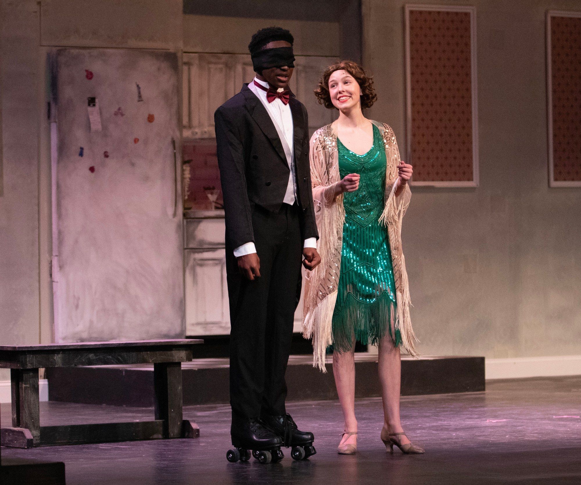 Amelie Lock in a green beaded 1920s dress and a fringed shawl, holding hands with a blindfolded performer on roller skates, in The Drowsy Chaperone