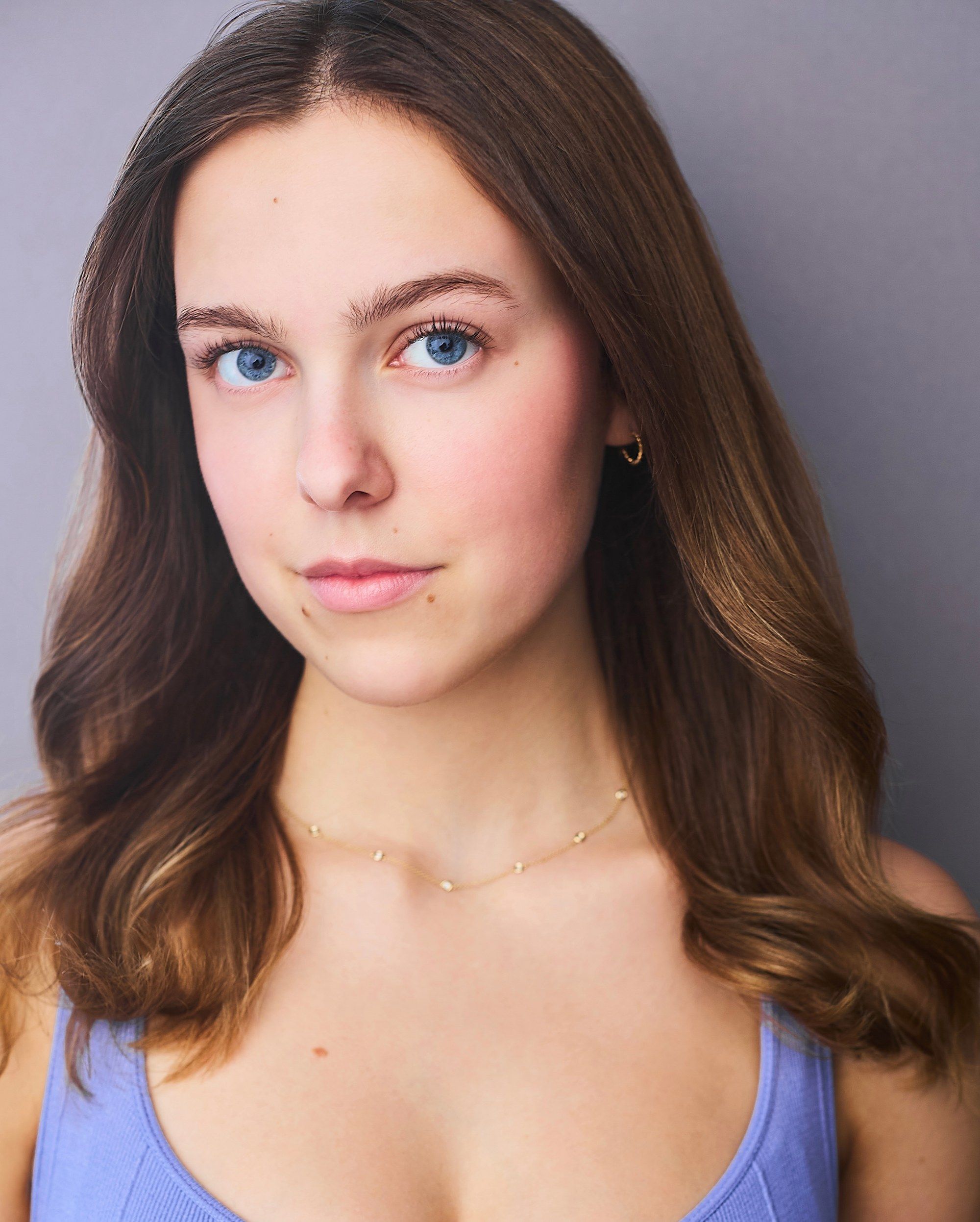 Amelie Lock, studio portrait in a lilac top and a thin gold necklace, long brown hair, blue eyes, a calm direct look, against a gray backdrop