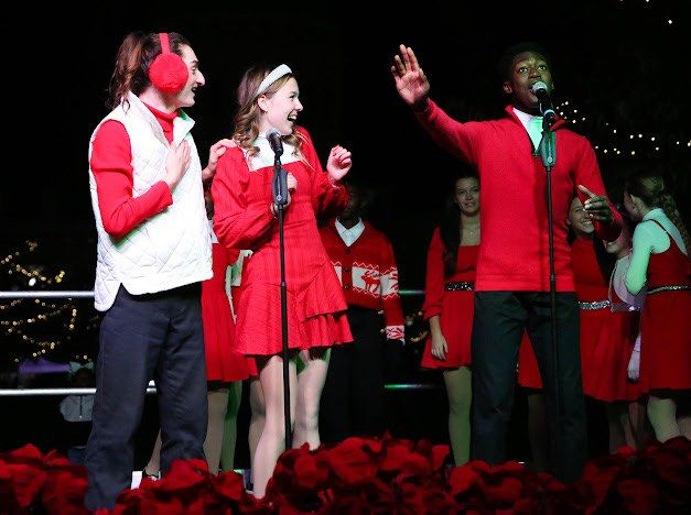 Performers in red and white winter outfits laughing and singing at a microphone in front of poinsettias and string lights