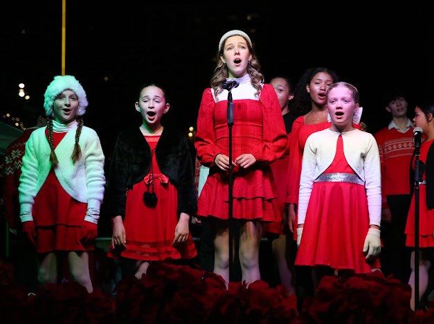 A chorus of children and teens in red outfits and winter hats singing into microphones in front of rows of poinsettias at night