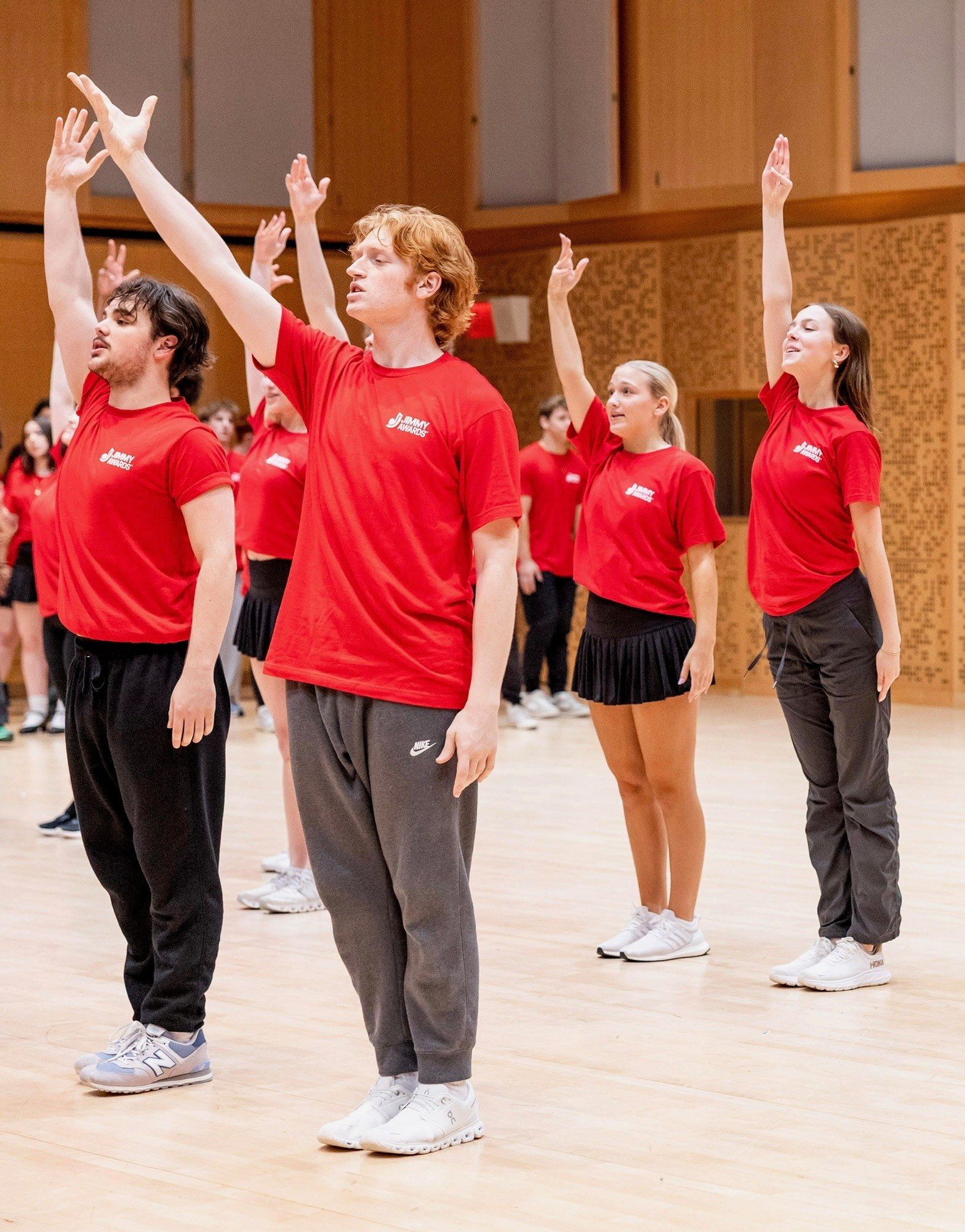 Jimmy Awards nominees in red T-shirts, arms raised high in a rehearsal studio