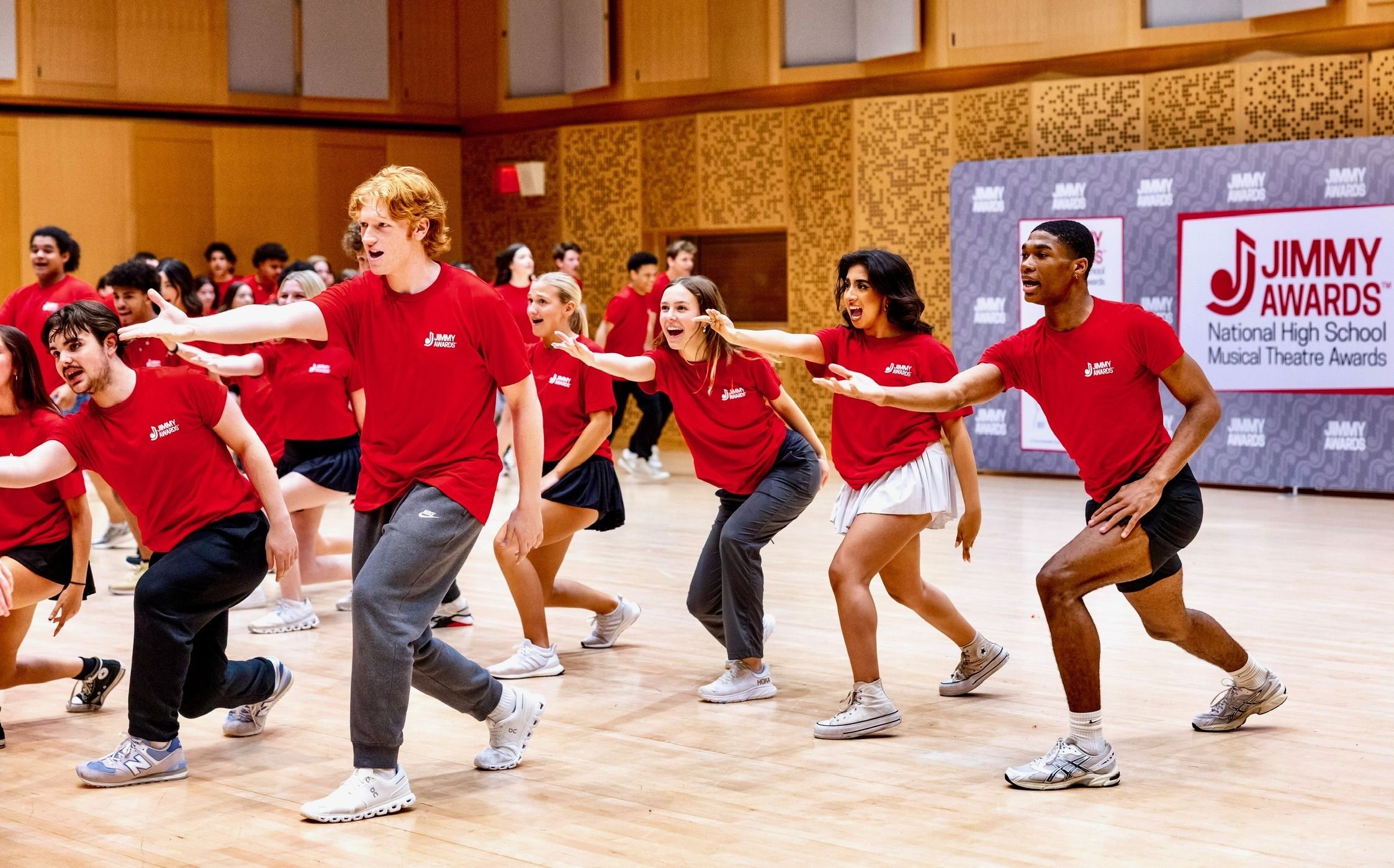 Jimmy Awards nominees in red T-shirts dancing across a wood-paneled rehearsal studio, arms outstretched