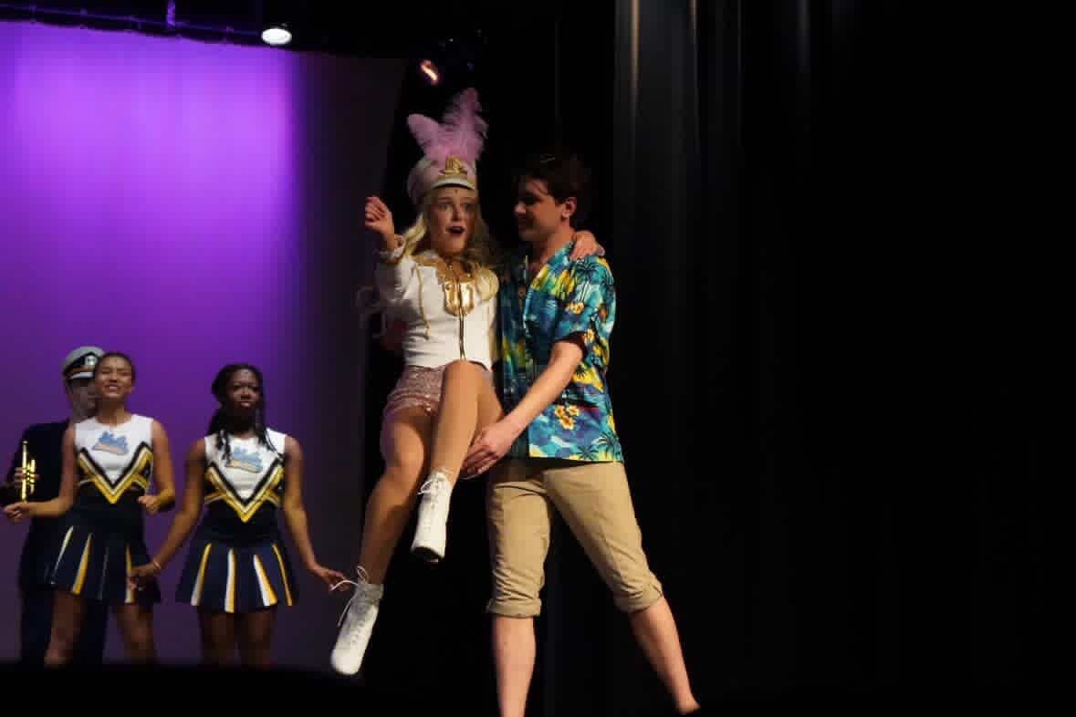 A performer in a white marching band uniform and feathered hat being lifted by a performer in a Hawaiian shirt, with cheerleaders behind them, in Legally Blonde