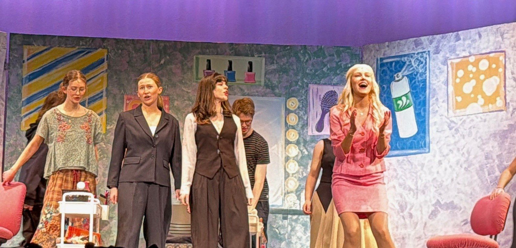 The cast on a painted salon set with nail polish and hairdryer artwork, one performer in a pink suit laughing and clapping at the front, in Legally Blonde