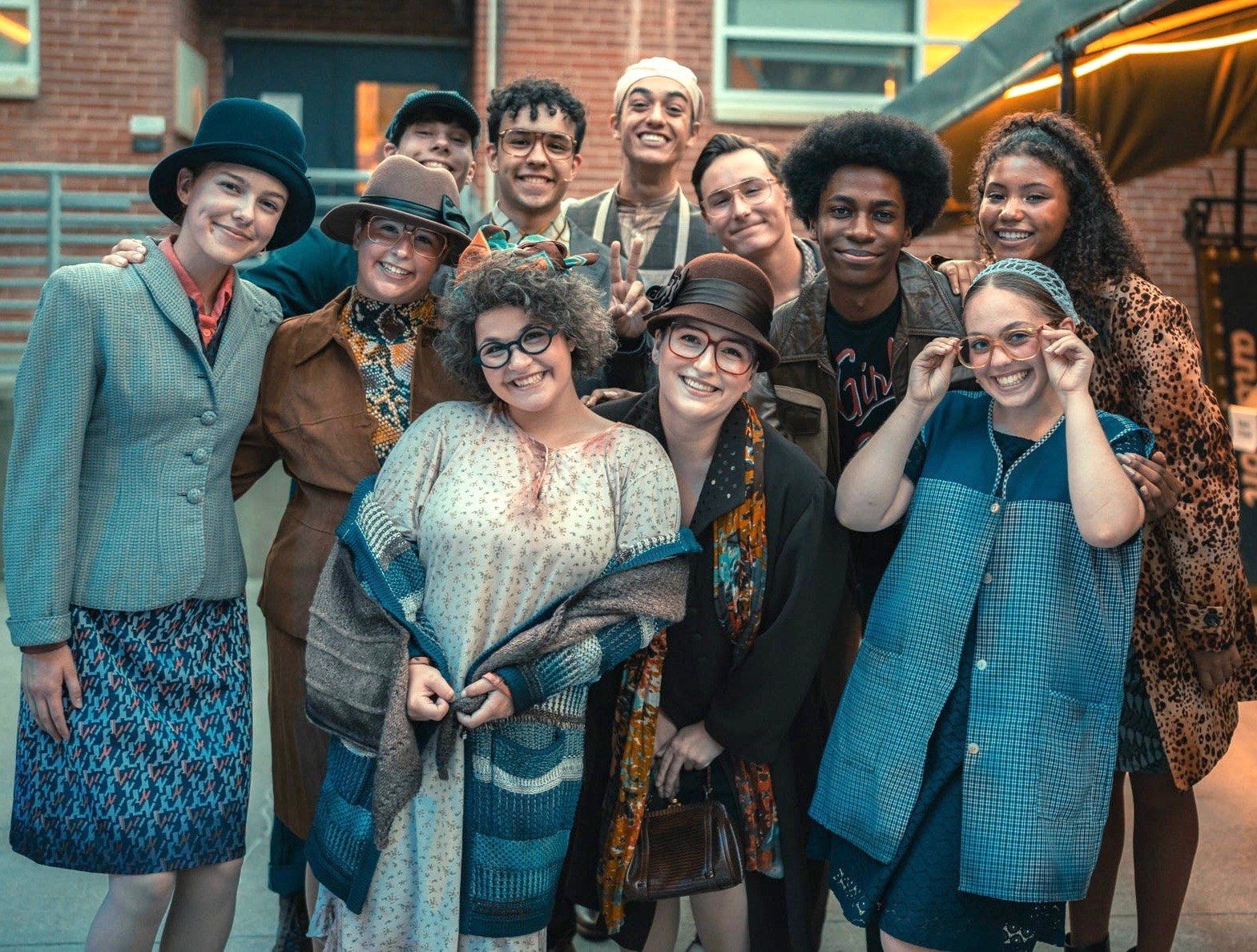 A smiling group of cast members in 1960s costumes, hats and glasses posed together outside a brick building, in Little Shop of Horrors at The Muny