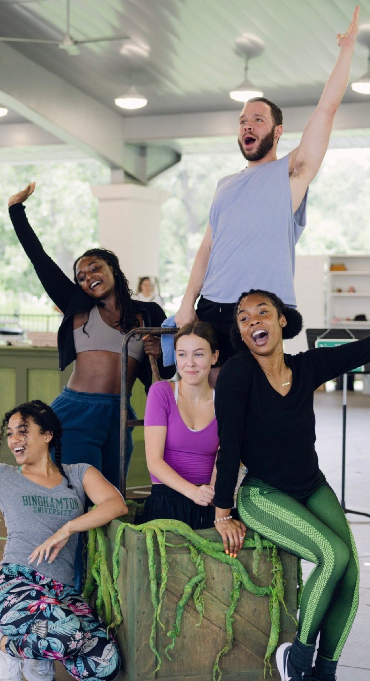 Five cast members in rehearsal clothes, laughing and posing with their arms in the air around a wooden crate with green tendrils, in a rehearsal pavilion for Little Shop of Horrors