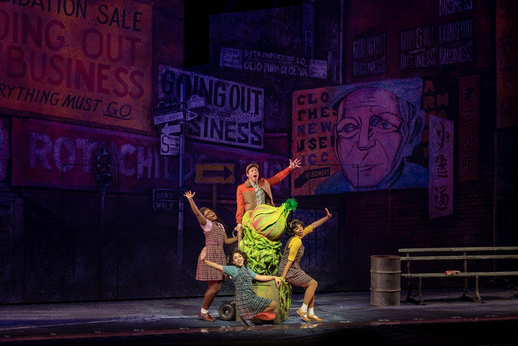 Four performers in 1960s costumes posed around the giant plant, Audrey II, in front of painted Going Out of Business signs in Little Shop of Horrors at The Muny