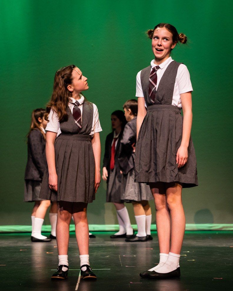 Two students in gray school pinafores on a green-lit stage, one looking up at the other, with classmates behind them, in Matilda