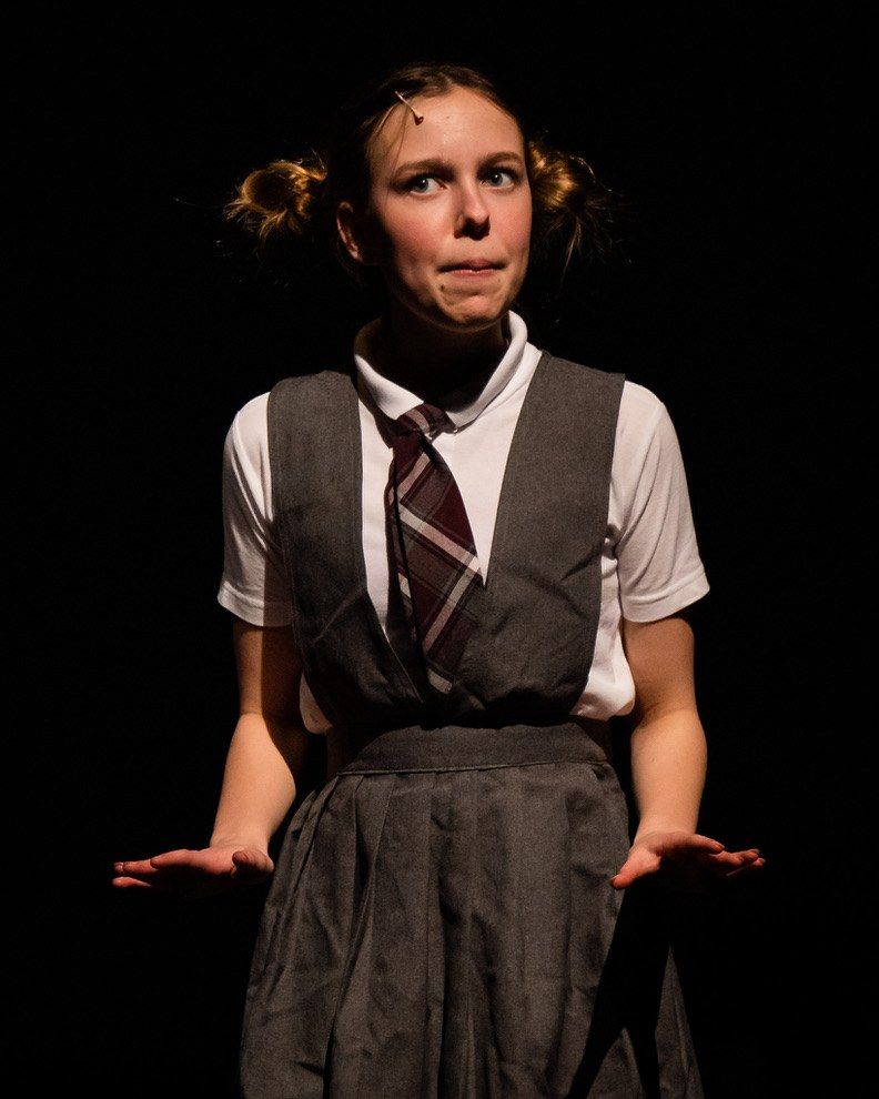 A performer in a gray school pinafore and striped tie, hair in two buns, lit alone against a black stage with a wry look, in Matilda