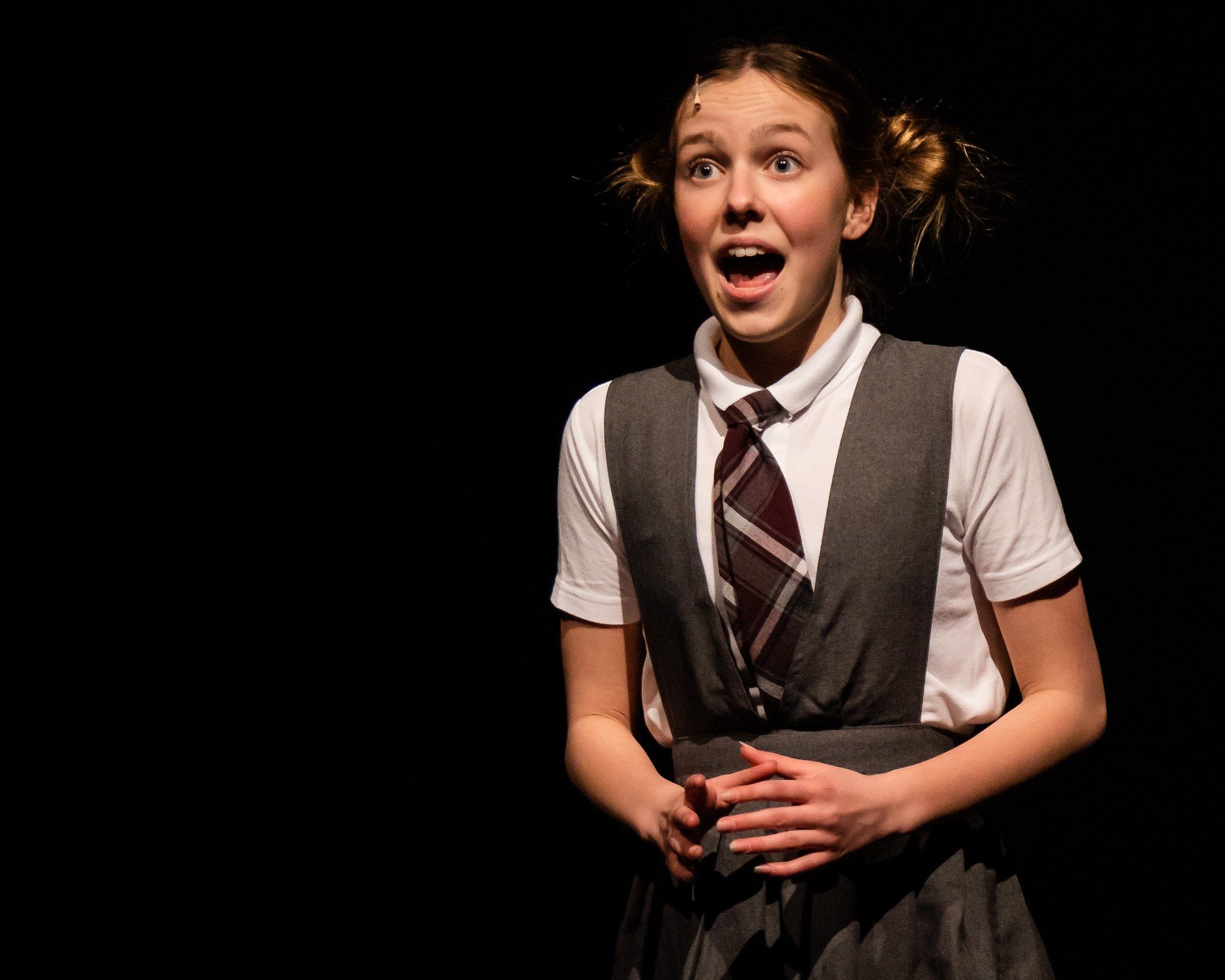 A performer in a gray school pinafore and striped tie, hair in two buns, caught mid-gasp against a black stage in Matilda