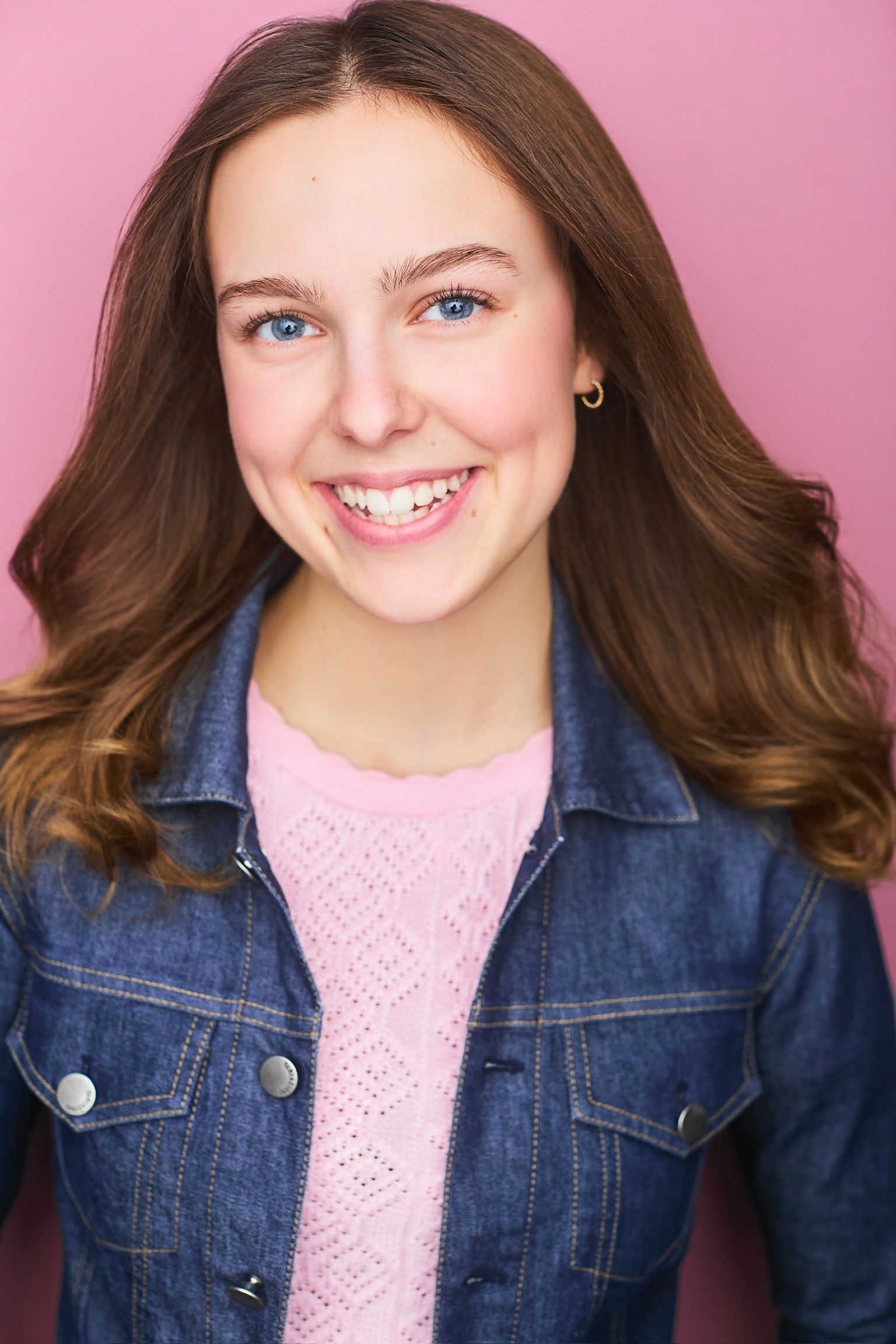 Amelie Lock smiling in a denim jacket over a pink knit top, against a pink studio backdrop