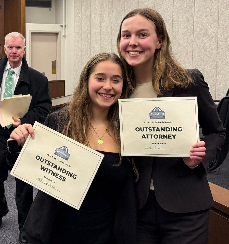 Two students in dark blazers smiling and holding printed mock trial award certificates in a courtroom