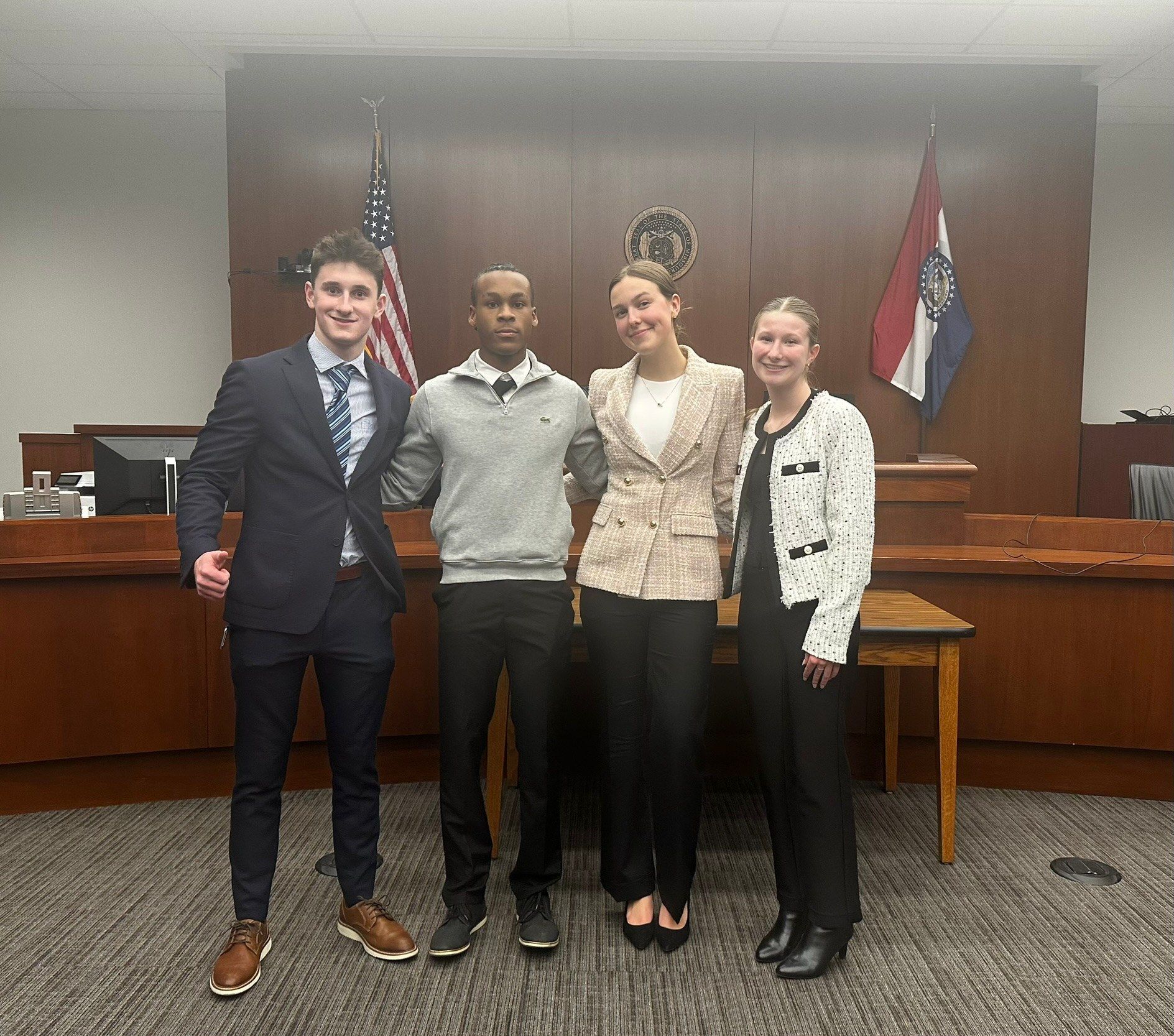 Four students in business clothes standing arm in arm in a courtroom, with American and Missouri flags behind them