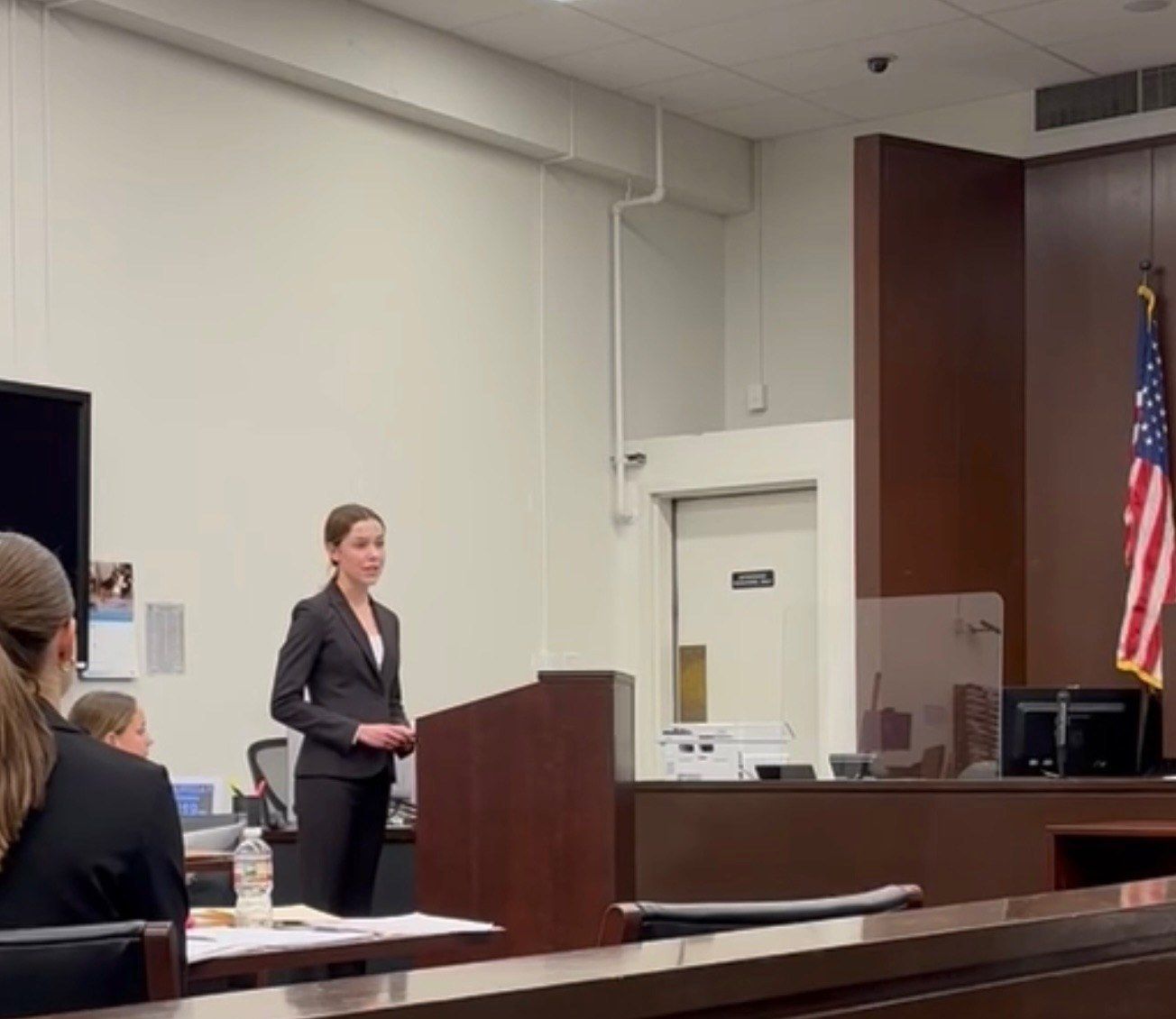 A student in a dark suit standing and speaking at a lectern in a courtroom, with an American flag nearby