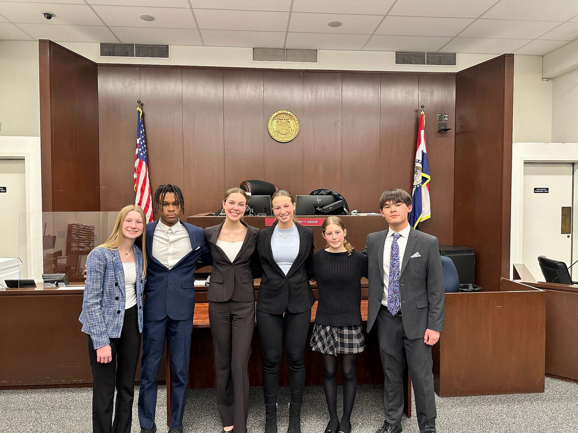 Six students in business clothes standing arm in arm in a courtroom in front of the judge’s bench