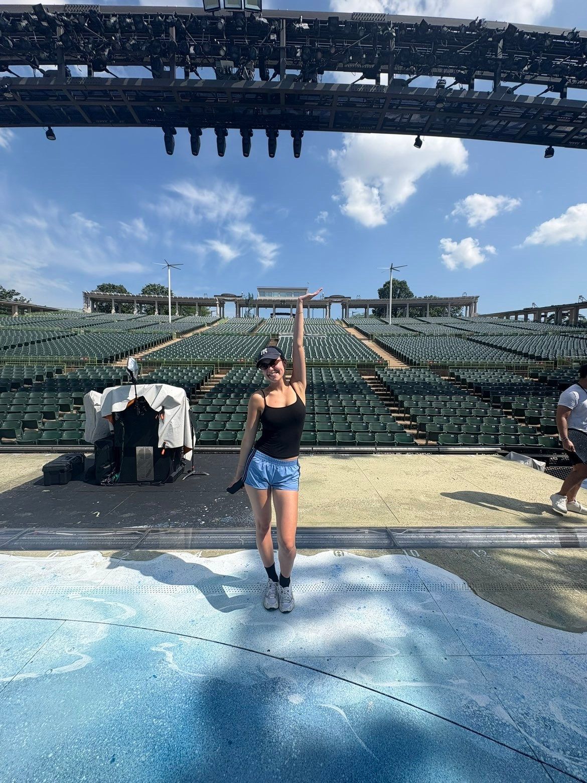Amelie Lock smiling in a cap and sunglasses, one arm raised, standing on the empty Muny stage with rows of green seats behind her