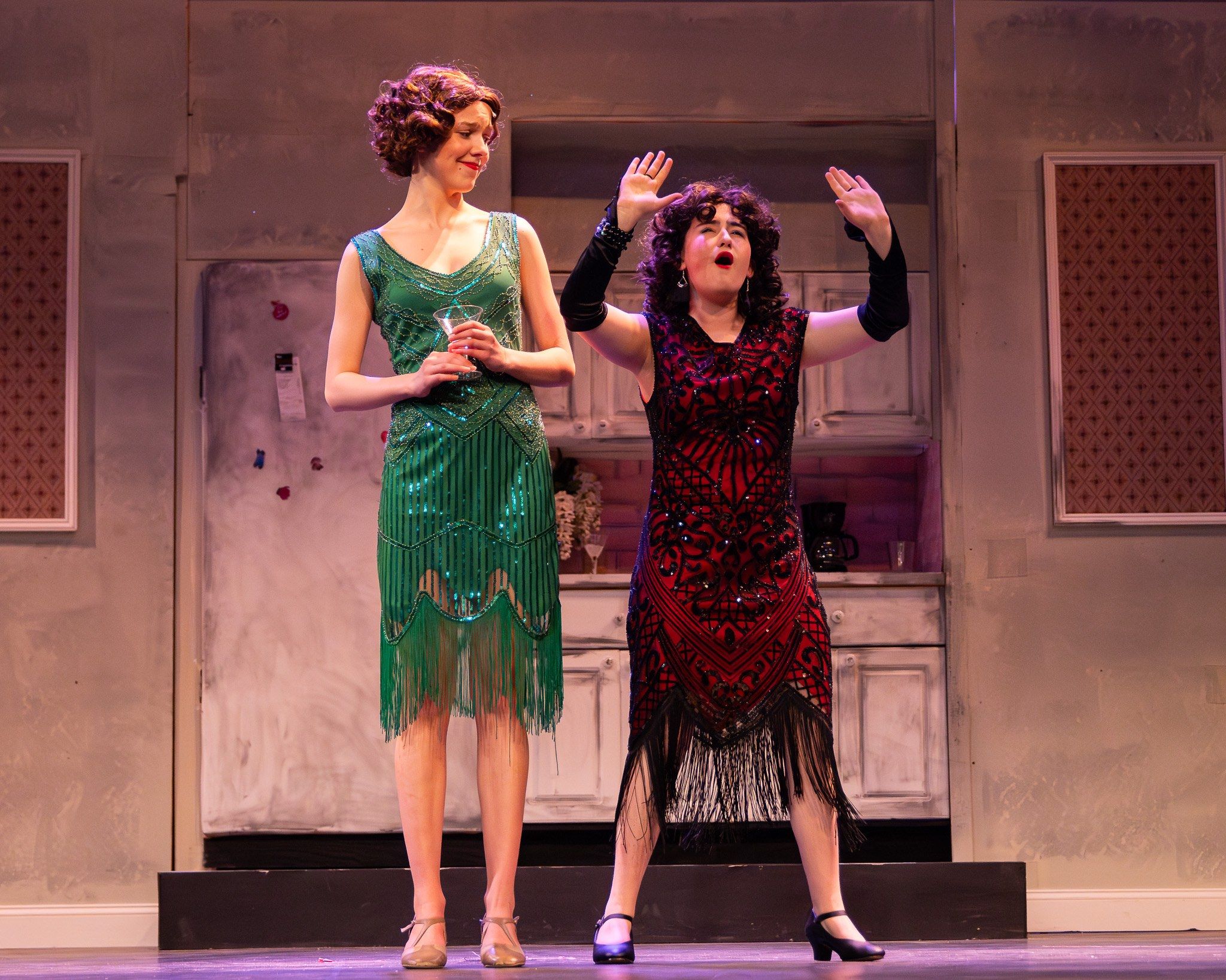 Two performers in 1920s dress, one in green beads and one in dark red, on a stage set with patterned panels, in The Drowsy Chaperone
