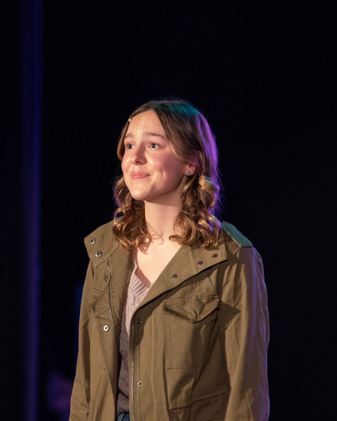 Amelie Lock on a dark stage in an olive utility jacket, a soft, hopeful look past the camera