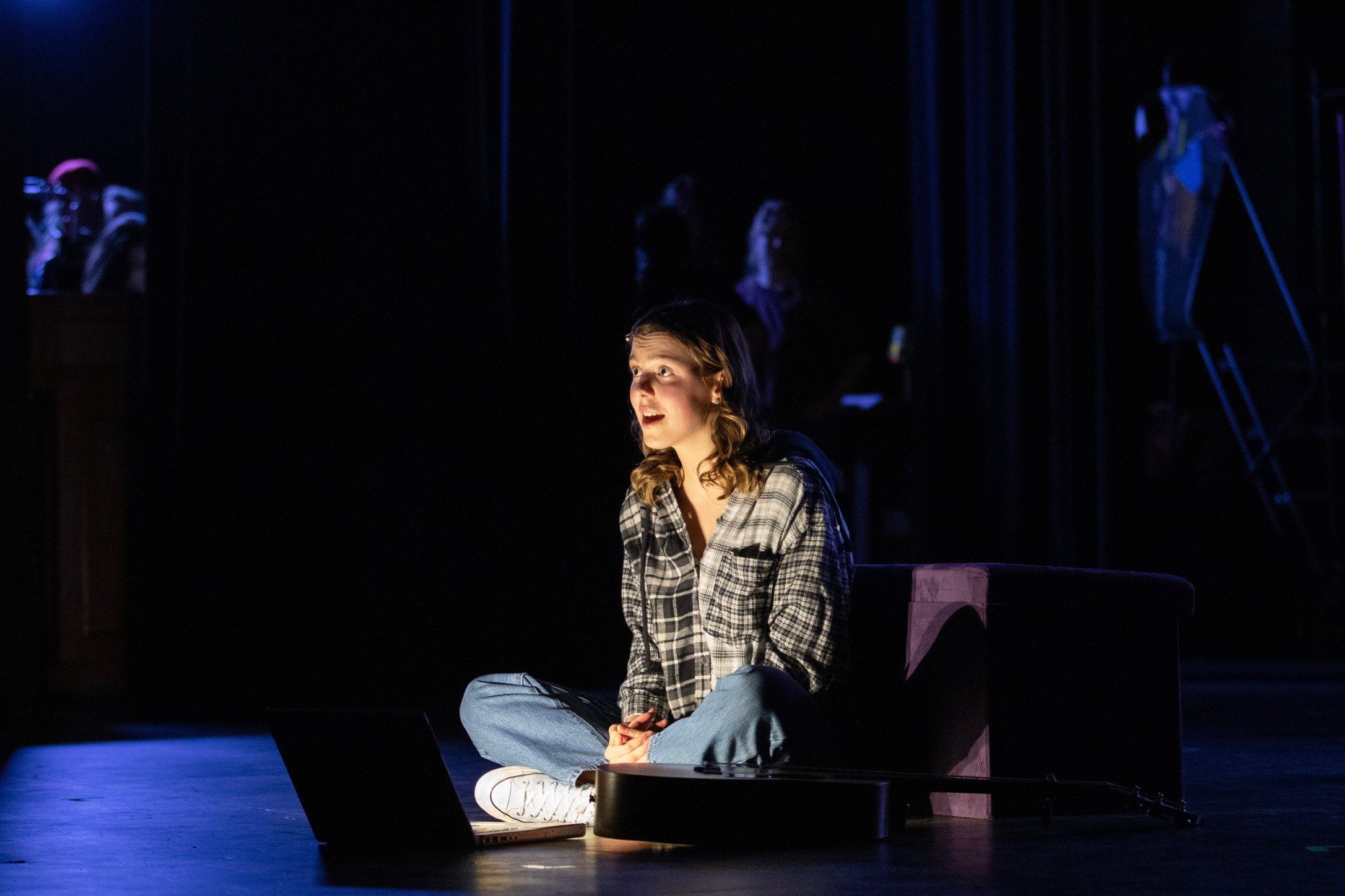 Amelie Lock sitting cross-legged on a dark stage in a plaid shirt, singing, a guitar beside her, in The Prom