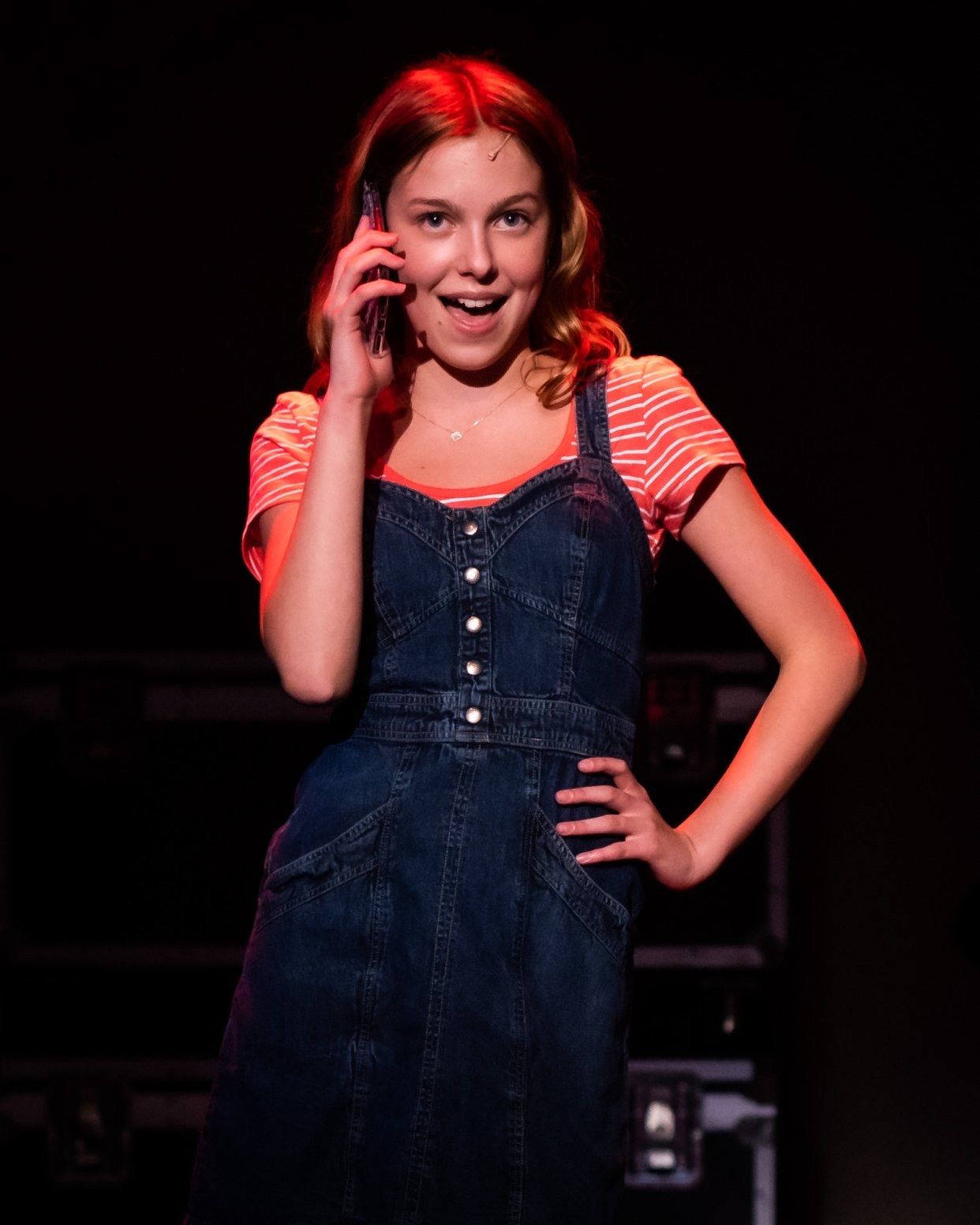 Amelie Lock as Lucy in denim overalls and a striped orange T-shirt, one hand to a phone at her ear and the other on her hip, lit in red against a dark stage, in 13: The Musical