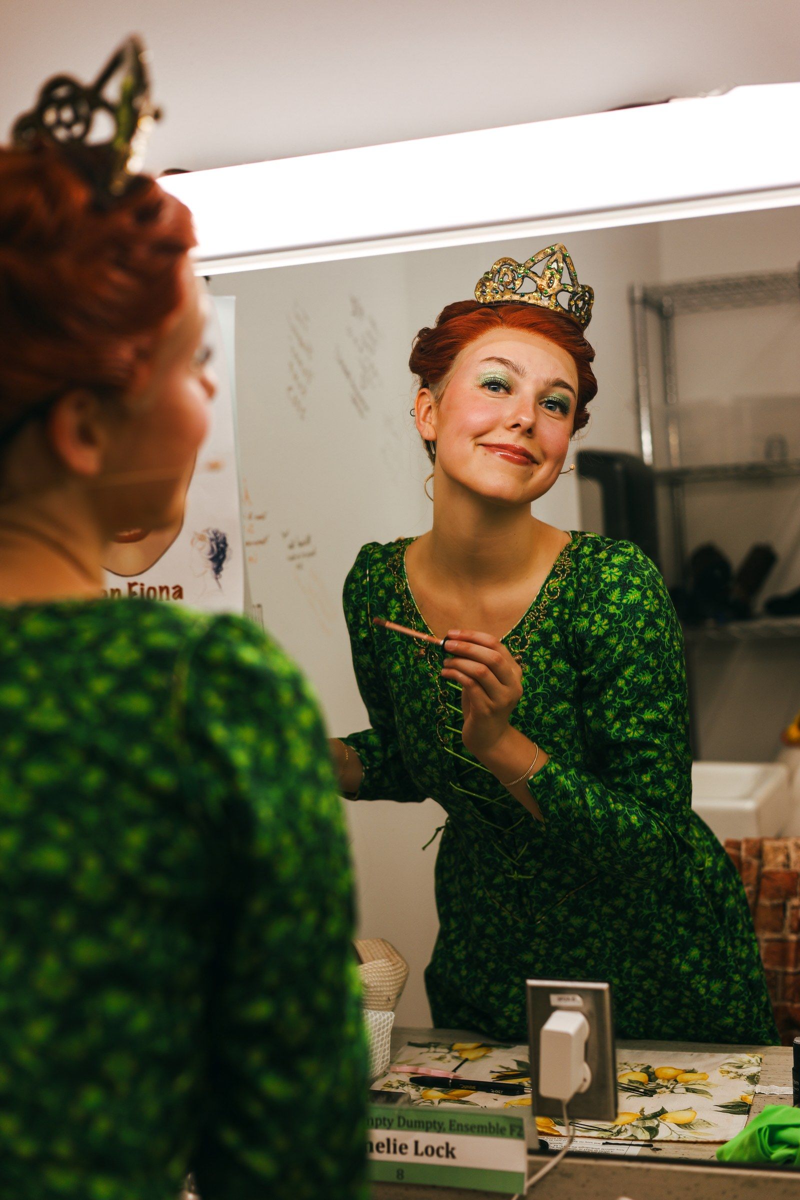 Amelie Lock in a green gown, red wig and crown, smiling at her reflection in a dressing room mirror as she applies makeup, backstage at Shrek The Musical