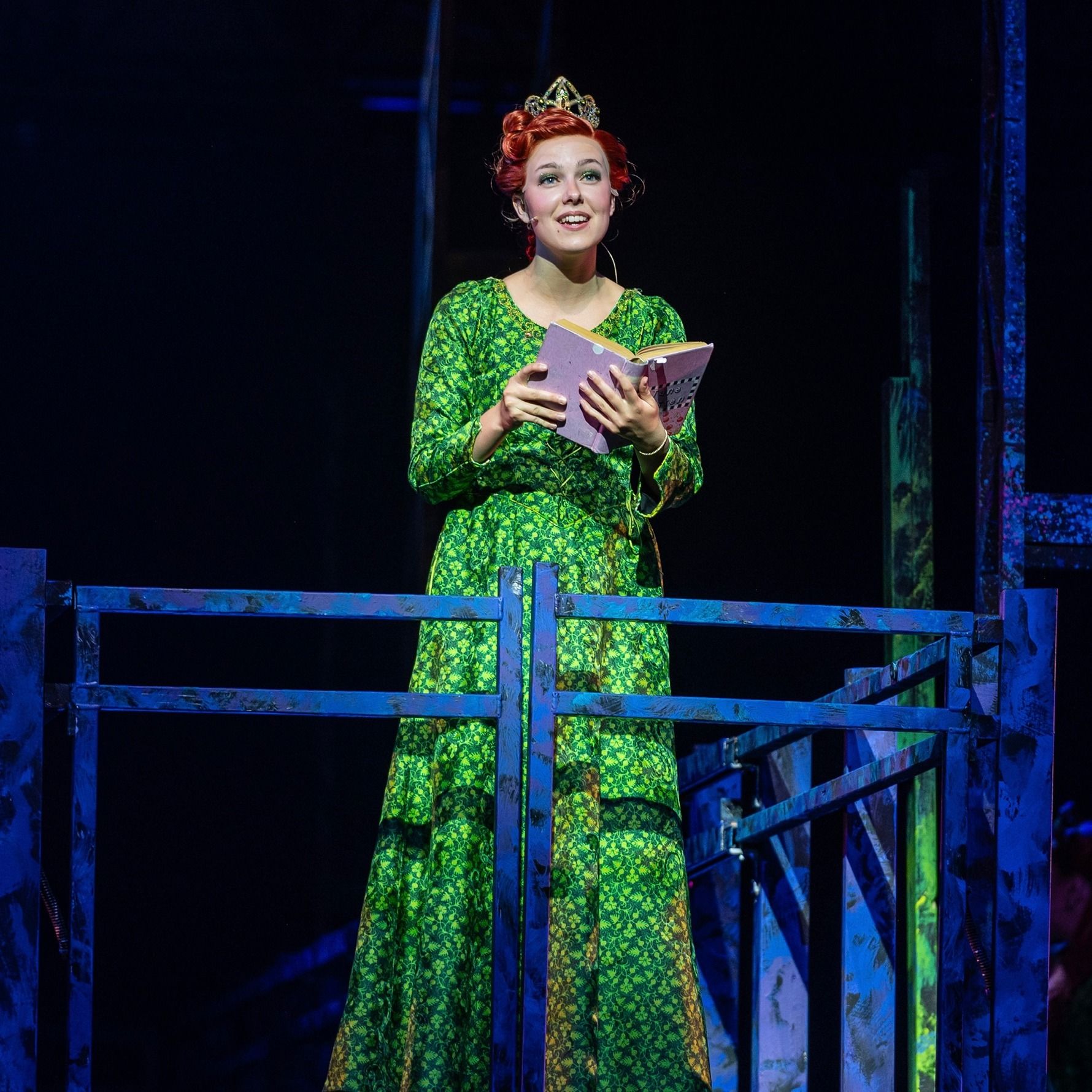 Amelie Lock as Teen Fiona in a green gown and crown, reading on the tower balcony in Shrek The Musical at The Muny