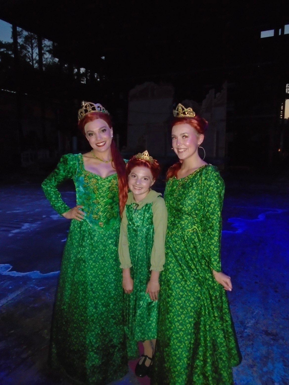 Three performers in green gowns, red wigs and crowns, one a young girl in the middle, posed together on the Muny stage after a performance of Shrek The Musical