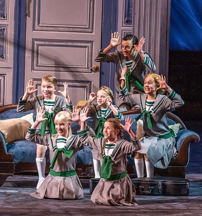 The von Trapp children in sailor costumes with their hands raised, posed around a blue sofa in The Sound of Music at The Muny