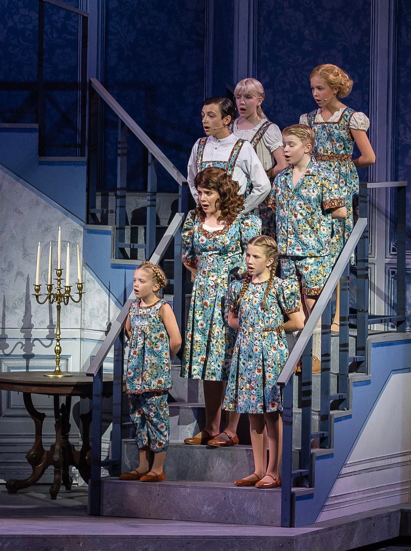 The von Trapp children in matching blue floral costumes standing in a line down a staircase, singing, in The Sound of Music at The Muny