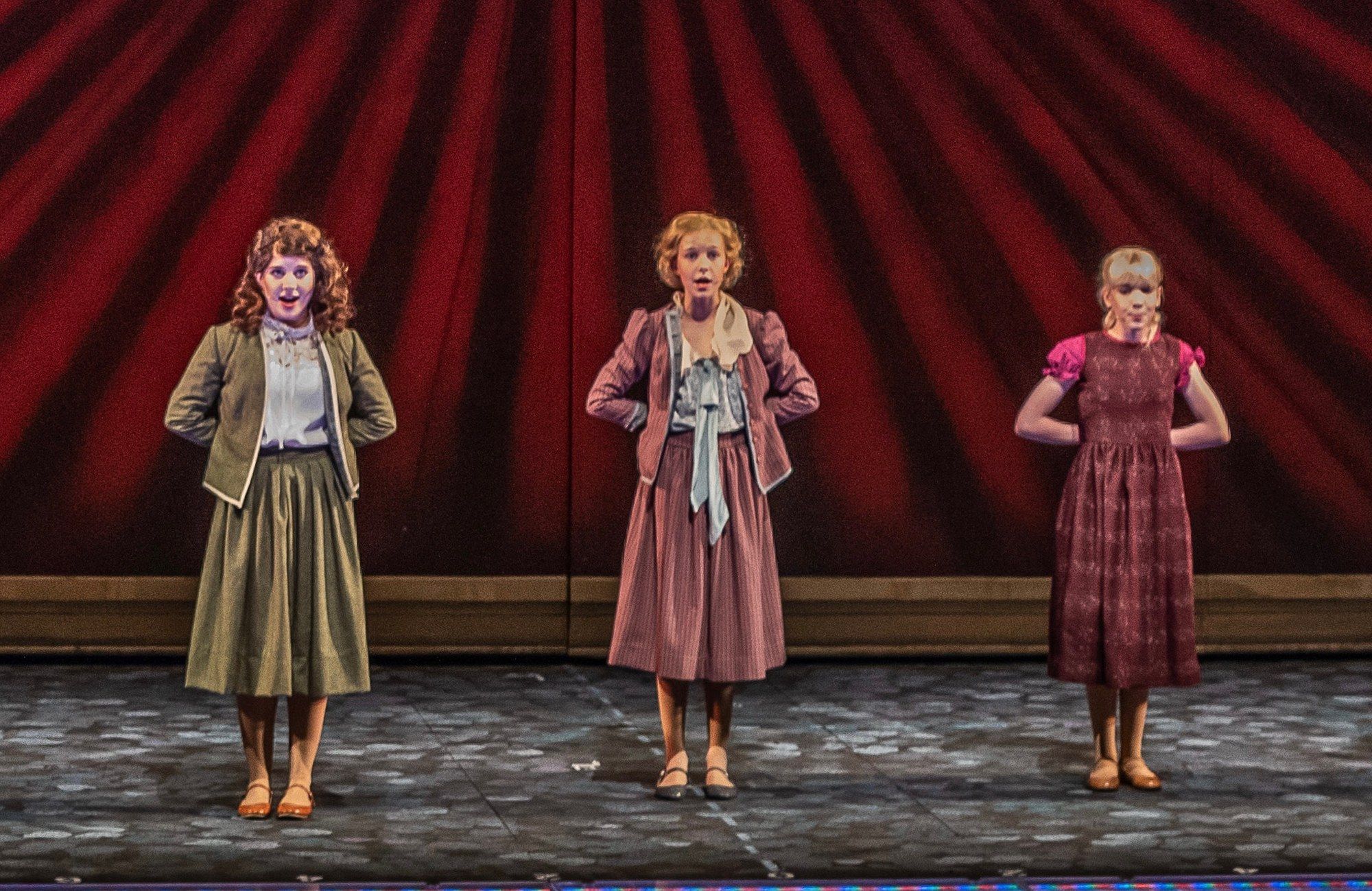 Three of the von Trapp children standing side by side with their hands behind their backs in front of a red sunburst backdrop, in The Sound of Music at The Muny