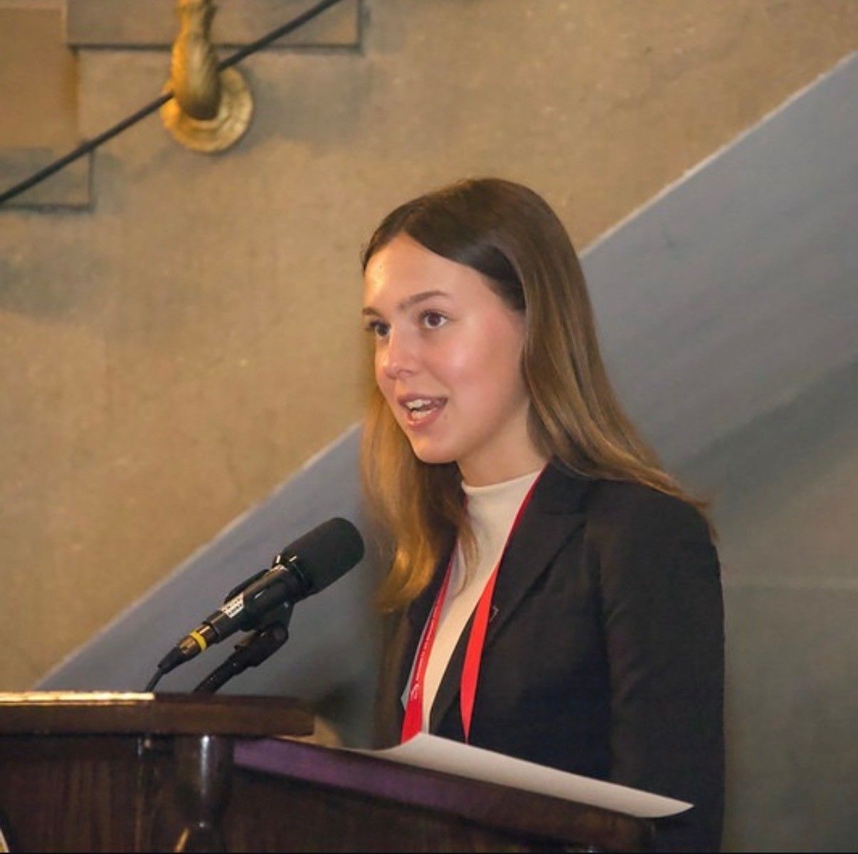 Amelie Lock speaking at a wooden podium with a microphone, in a dark blazer with a red lanyard