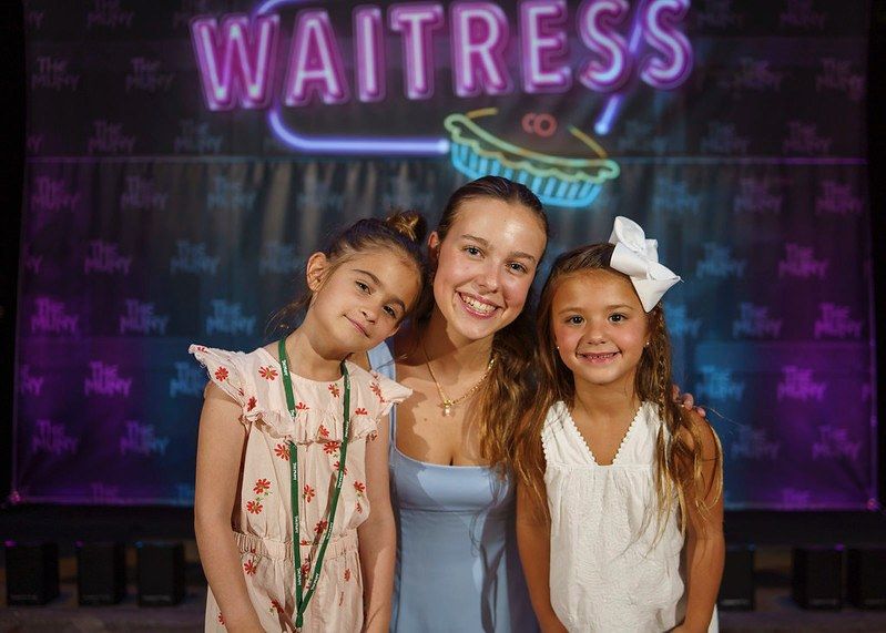A smiling young woman between two little girls in front of a neon Waitress sign backdrop at The Muny