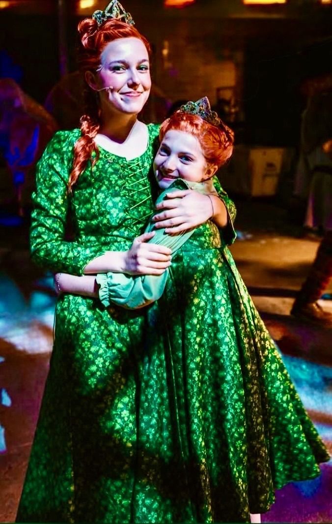 Two performers in green gowns and crowns embracing, in Shrek The Musical at The Muny